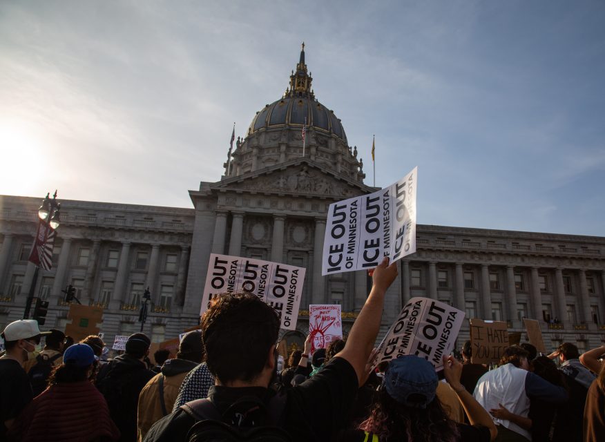 A crowd of protesters holds signs reading “ICE OUT OF MINNESOTA” in front of a large government building with a domed roof.