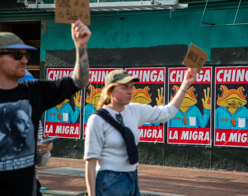 Two people hold protest signs in front of a wall featuring repeated posters with a cartoon frog and the words "CHINGA LA MIGRA.