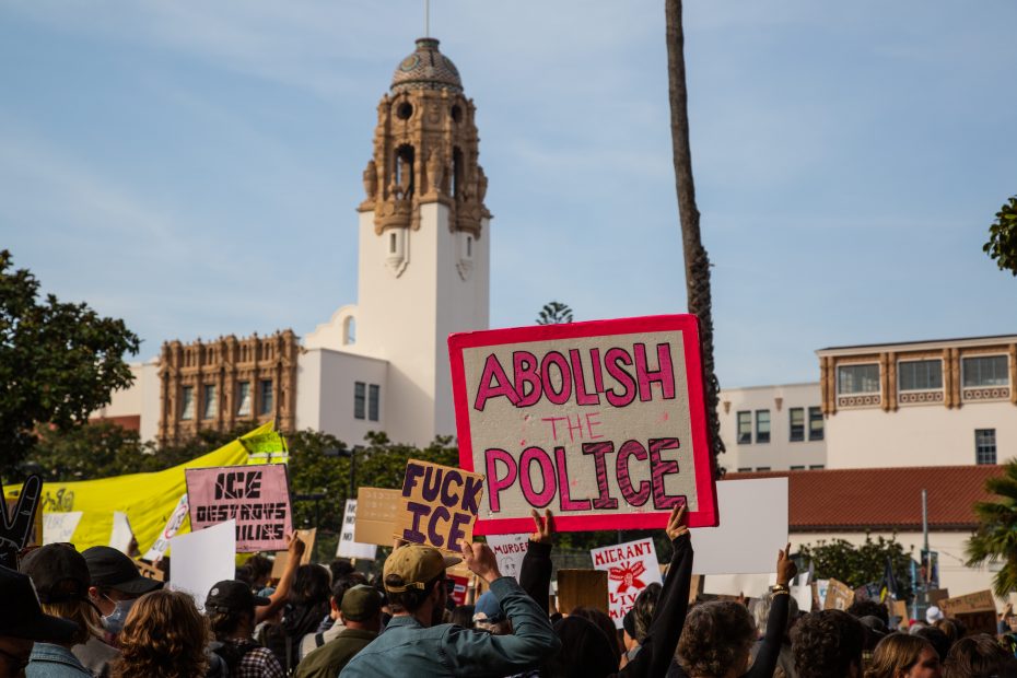 A crowd of protesters holds signs, including one reading "Abolish the Police," in front of a historic building with a tower and palm trees.