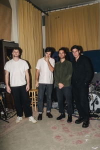 Four men stand indoors next to musical equipment and drums, in front of a curtain. They face the camera, wearing casual clothing.
