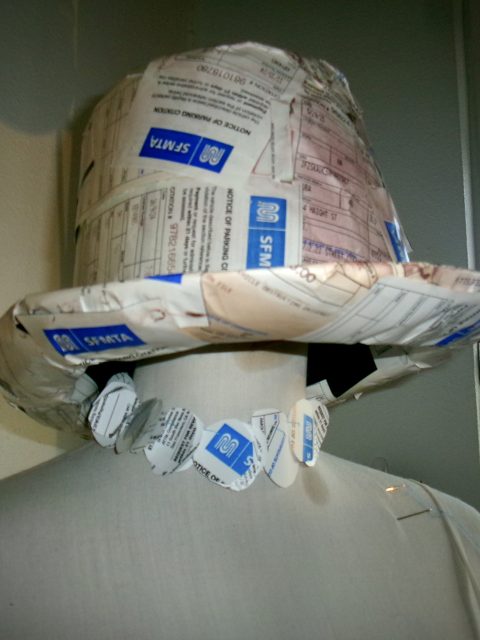 A mannequin wearing a hat and necklace made from folded and cut paper, including parking tickets and forms with blue and white text.