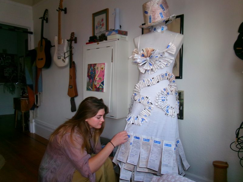 A woman kneels beside a mannequin dressed in a white outfit decorated with paper parking tickets, in a room with guitars hanging on the wall.