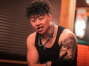 A man with a chain necklace and a tattooed arm wears a black tank top and poses indoors, looking toward the camera.