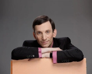 A man in a suit with a pink shirt leans his arms on a peach-colored surface, looking directly at the camera against a plain gray background.