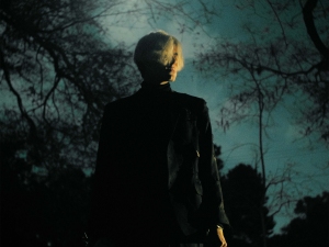 A person with short light hair stands outside at night, silhouetted against a cloudy, dimly lit sky with bare tree branches.