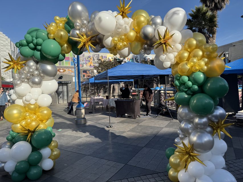 A decorative arch made of green, white, gold, and silver balloons with gold star accents stands in front of blue canopy tents in an outdoor plaza.