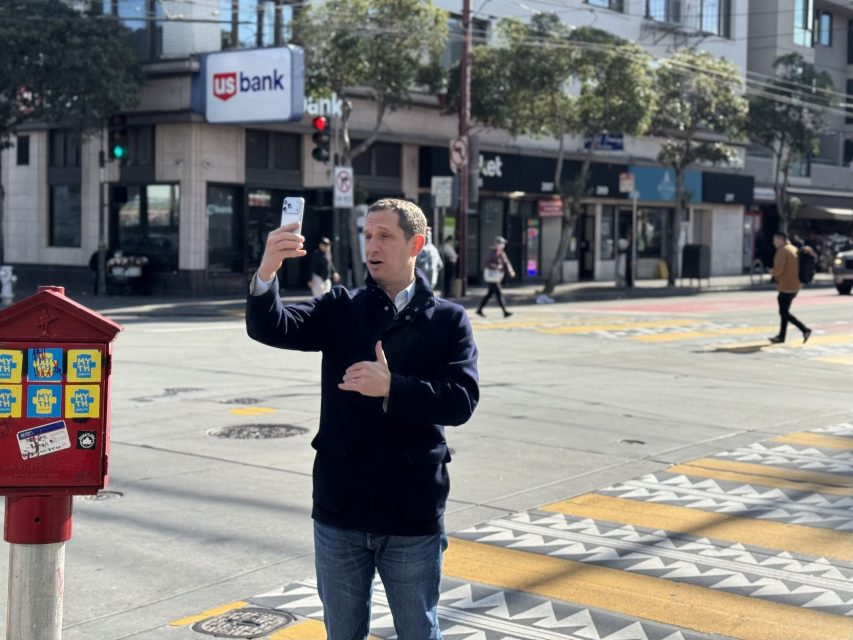 A man stands on a city street corner holding a smartphone, appearing to record himself. Buildings, crosswalks, and a US Bank branch are visible in the background.