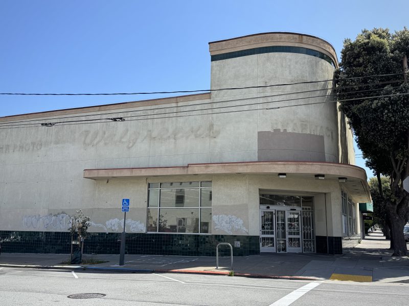 A vacant corner storefront with faded signage, barred front doors, and green tile accents along the lower wall on a sunny day.