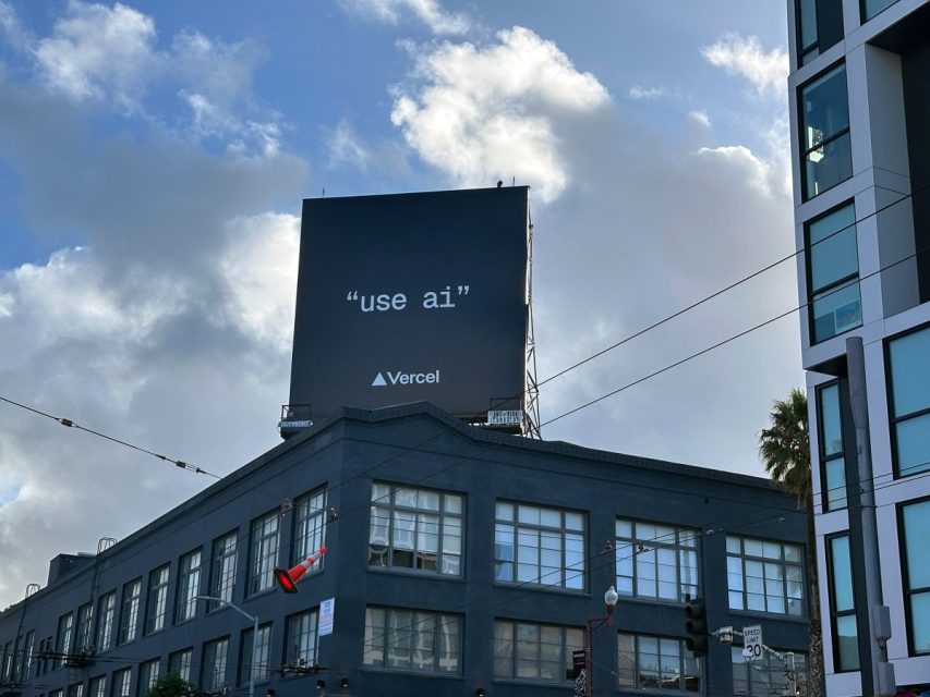 A black billboard on top of a building displays the text “use ai” and the Vercel logo against a partly cloudy sky.
