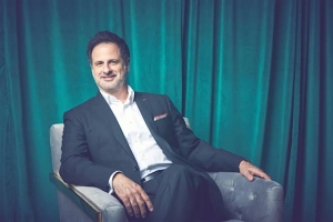 A man in a dark suit sits in a gray armchair in front of teal curtains, looking at the camera and smiling slightly.
