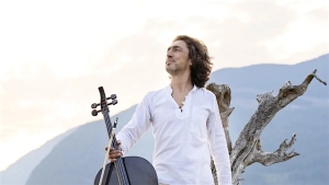 A person in a white shirt stands outdoors holding a cello, with mountains and a tree in the background.