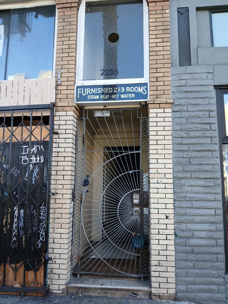 Narrow entrance to a brick building with a decorative spiral metal gate, a sign reading "Furnished Rooms Steam Heat Hot Water," and the street number 2230 above the door.