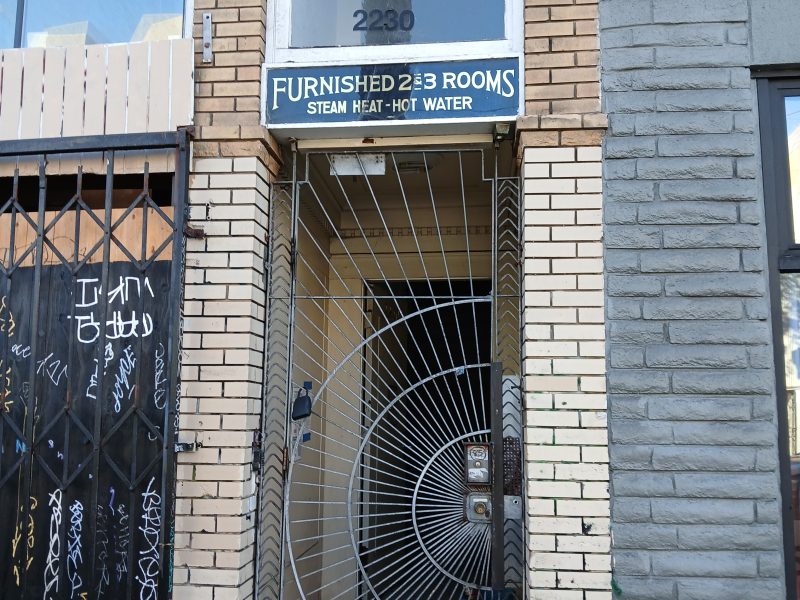 Narrow entrance to a brick building with a decorative spiral metal gate, a sign reading "Furnished Rooms Steam Heat Hot Water," and the street number 2230 above the door.