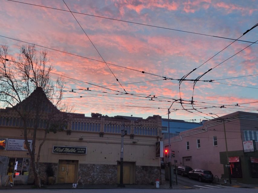 Street scene at sunset showing a building with closed doors, overhead trolley wires, a bare tree, and a sky filled with pink clouds.