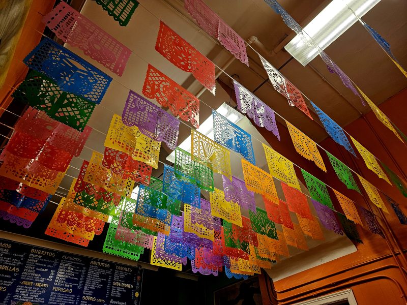 Colorful papel picado banners hang from the ceiling inside a building, with a menu board visible on the wall below.