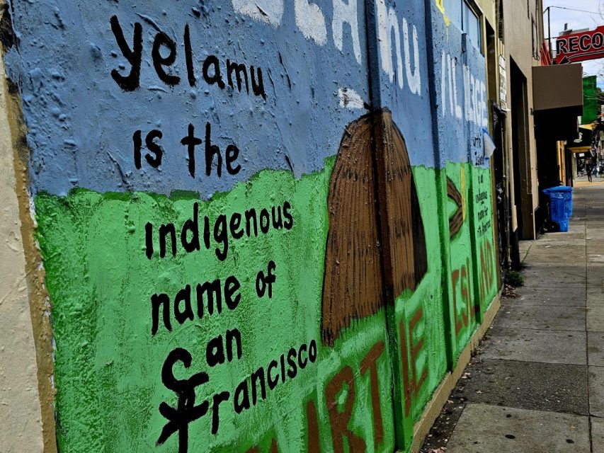 A mural on a city sidewalk wall reads, "Yelamu is the indigenous name of San Francisco," with colorful text and a dome-shaped hut illustration.