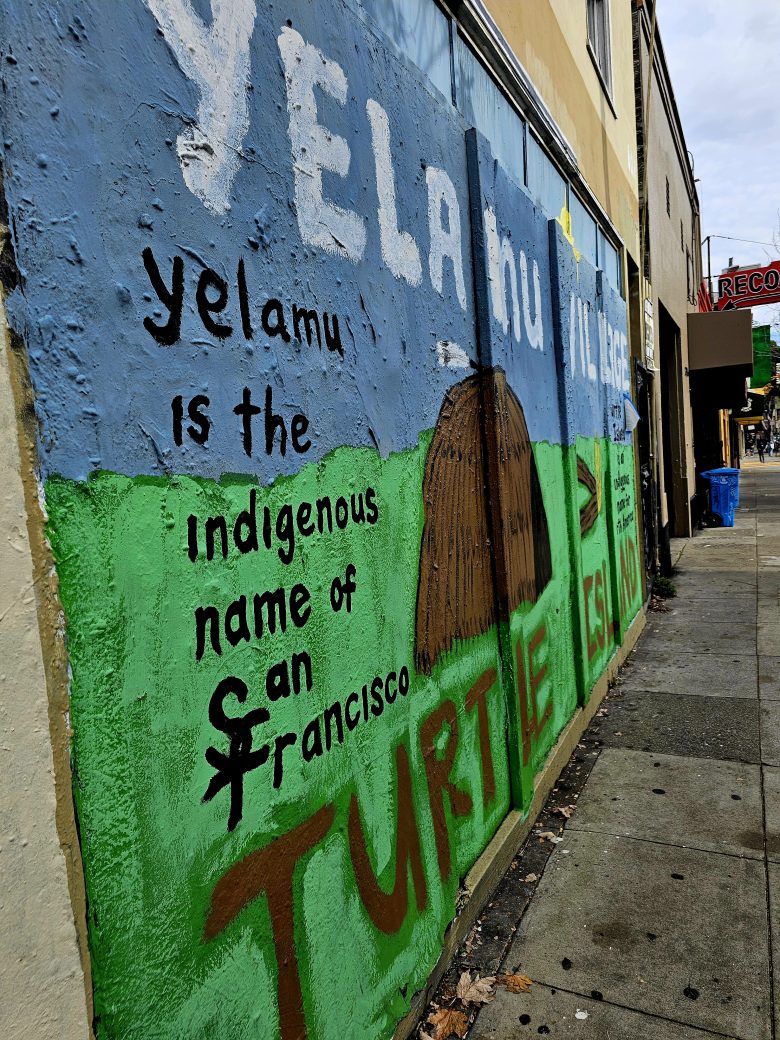 A mural on a city wall states, "Yelamu is the indigenous name of San Francisco," with colorful lettering and a hut illustration.