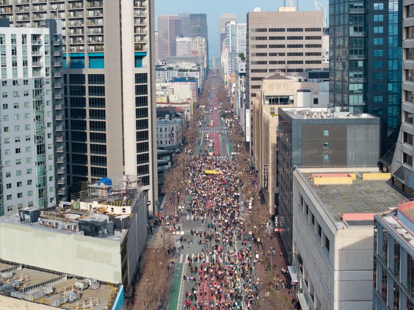 A large crowd fills a city street lined with tall buildings, seen from above, as an ICE walkout draws people together with more skyscrapers in the background.