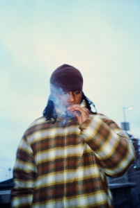 A person wearing a plaid jacket and beanie is standing outdoors, holding and smoking a cigarette with smoke partially obscuring their face.