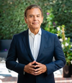 A man in a navy suit and white shirt stands outdoors with his hands clasped, green foliage and potted plants in the background.