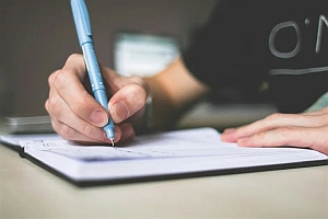 A person writes in a notebook with a blue pen. The focus is on the hand and pen, while the background is blurred.