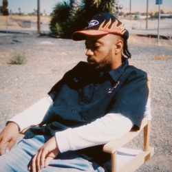 A man wearing a flame-patterned cap and dark shirt sits in a wooden chair outdoors on a gravel surface, with sunlight casting shadows.