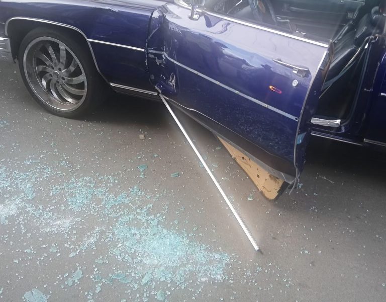 A blue car with a damaged door and shattered glass on the ground; the door is ajar and a metal rod lies beside it. People and barriers are visible in the background.