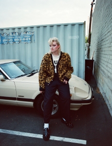 Person with short blonde hair wearing a leopard print jacket and black pants leans against a cream-colored vintage car in a parking lot next to a corrugated metal container.