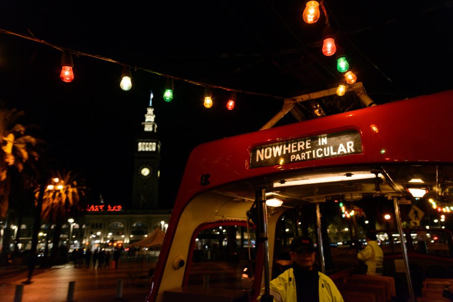 A red trolley with a sign reading "Nowhere in Particular" is parked at night under string lights, with a clock tower and city lights visible in the background.
