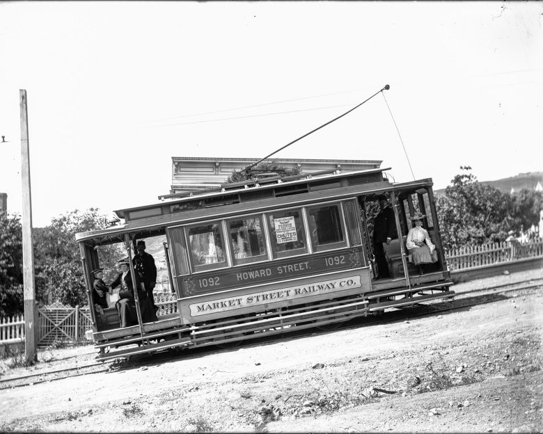Historic streetcar labeled "Howard Street 1092" of the Market Street Railway Co. tilted on a dirt road with passengers and conductor visible.
