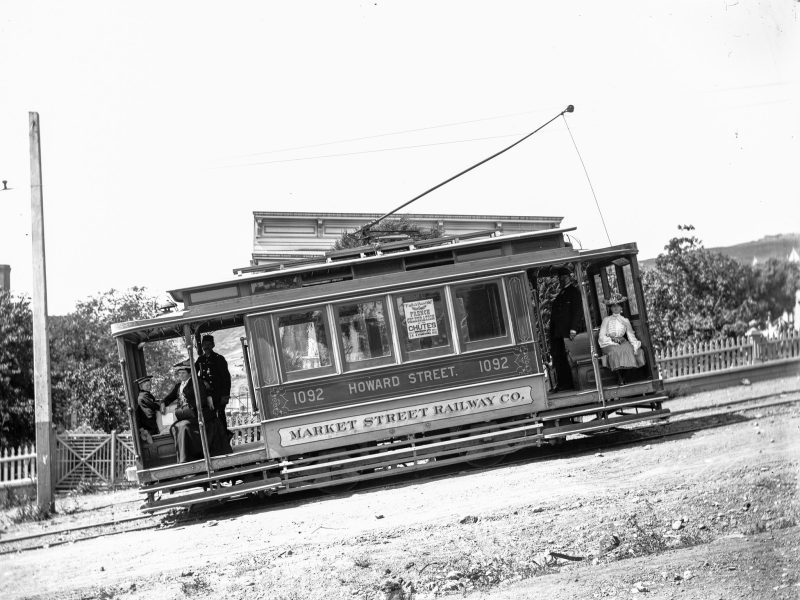 Historic streetcar labeled "Howard Street 1092" of the Market Street Railway Co. tilted on a dirt road with passengers and conductor visible.