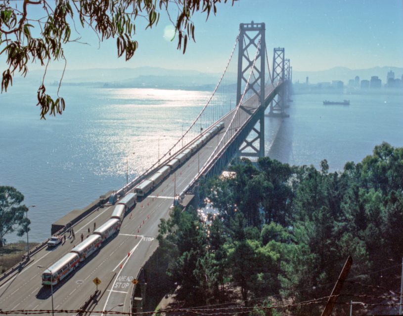 A line of buses crosses the San Francisco–Oakland Bay Bridge over calm water, with the city skyline and distant hills visible through light haze.