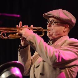 A man wearing glasses and a flat cap plays a trumpet on stage under pink and yellow lighting.