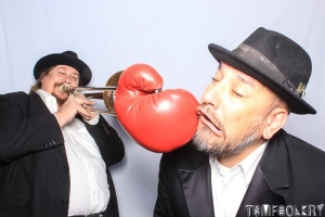 A man in a suit and hat is hit in the face with a red boxing glove while another man in the background plays a trumpet, both wearing black hats and jackets.