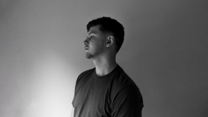 A young man with short hair and a beard stands in profile against a plain, light-colored background, looking slightly upward. The image is in black and white.