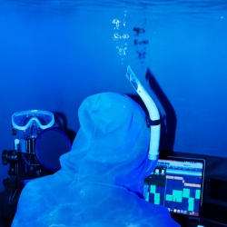 A person in blue snorkel gear uses a laptop underwater, with audio equipment and a microphone nearby.