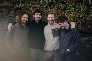 Four men stand close together smiling, with arms around each other, in front of a stone wall covered in ivy.