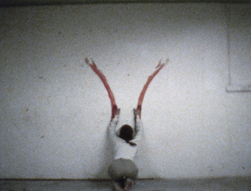 A person kneels facing a wall, extending both arms upwards to create two long red paint streaks on the white surface, evoking performance art scenes often found in contemporary museums.