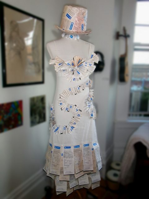 A mannequin displays a white dress and hat adorned with numerous New York City MTA MetroCards, parking tickets, and what appear to be transit tickets or receipts, set in an indoor space.