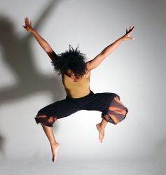A person with curly hair jumps in the air with arms and legs spread wide, casting a shadow on a plain background.