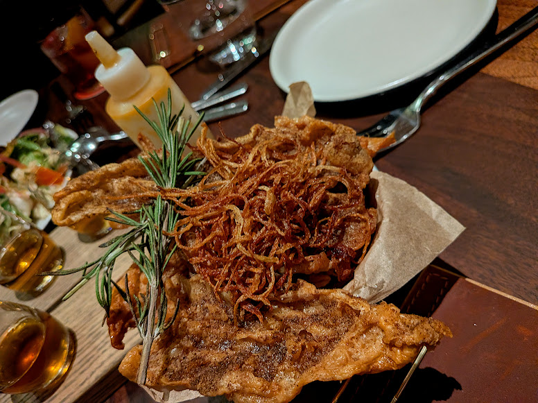 A close-up of a plate with fried food garnished with crispy onions and a sprig of rosemary, set on a wooden table beside an empty plate and utensils.