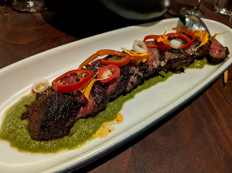 A grilled strip of steak served on green sauce, topped with sliced red peppers, onions, and edible flower petals on a white oval plate.