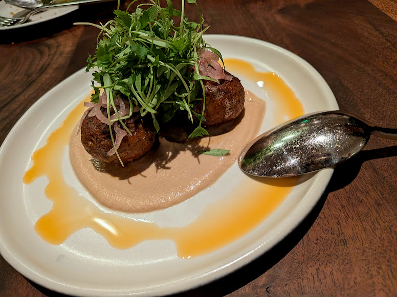 Two meatballs topped with greens and pickled onions on a layer of creamy sauce, garnished with oil, served on a white plate with a spoon.
