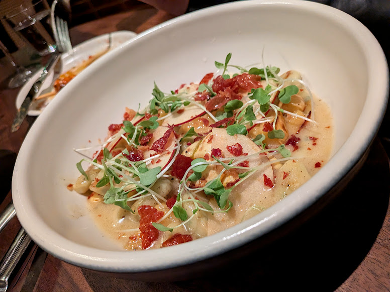 A white bowl filled with pasta in a creamy sauce, topped with microgreens and diced sun-dried tomatoes, sits on a wooden table.