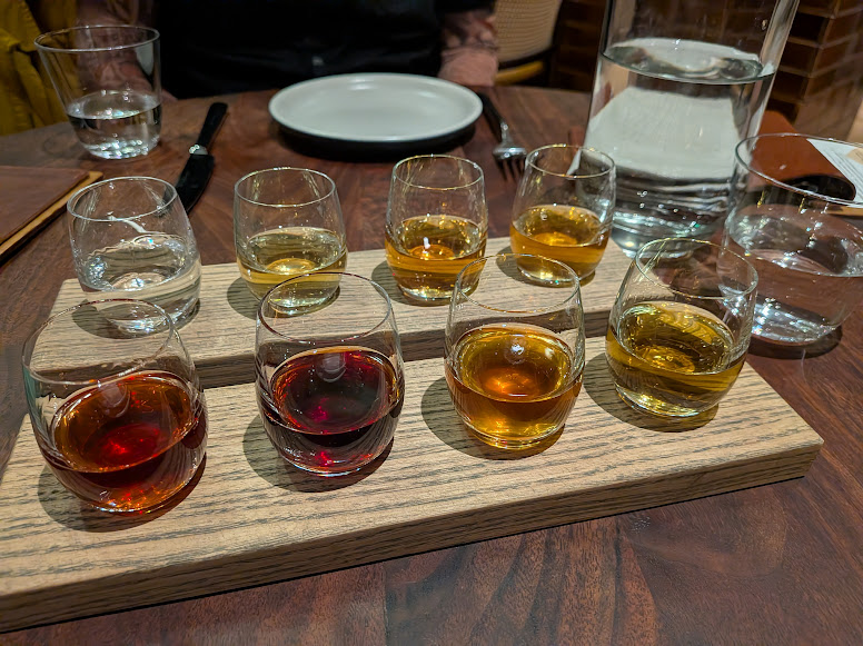 A wooden tray holds eight small glasses with different colored liquids, likely a tasting flight, on a restaurant table with plates and water glasses.