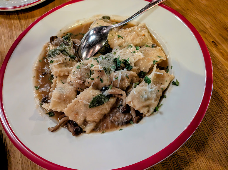 A plate of ravioli in a brown mushroom sauce, garnished with grated cheese and herbs, with a spoon on the side.