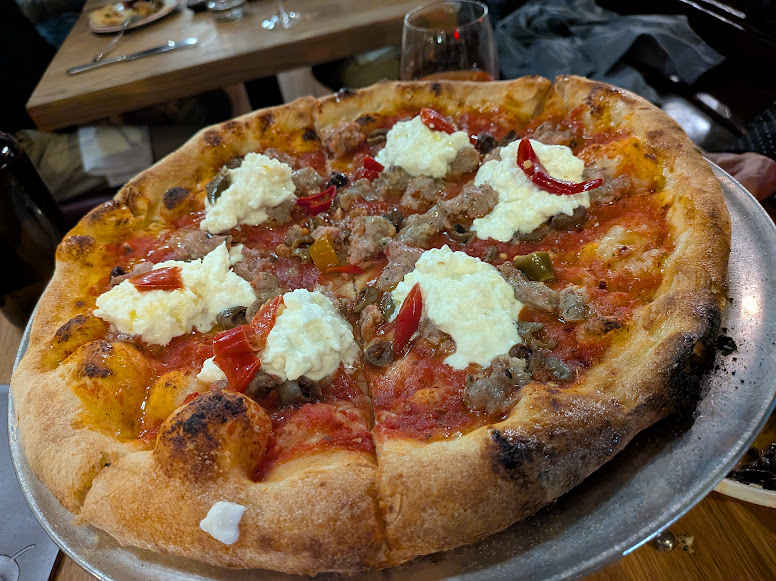A pizza topped with dollops of ricotta cheese, sausage, red pepper slices, and tomato sauce on a metal tray in a restaurant setting.