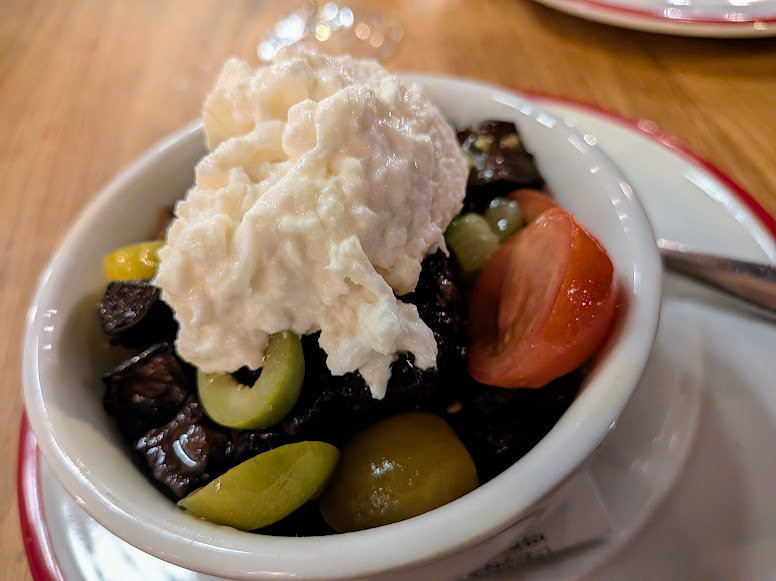 A white bowl contains olives, tomatoes, dark eggplant pieces, and a large dollop of ricotta cheese, placed on a white plate with a spoon beside it.