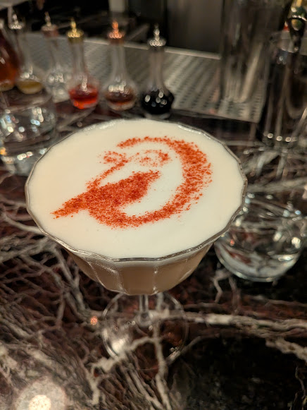 A cocktail with a foamy top, garnished with red powder in a swirl pattern, sits on a marble bar counter next to an empty glass and small bottles of ingredients.