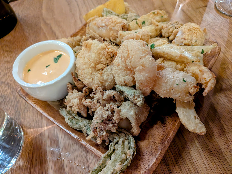 A wooden plate with fried seafood and vegetables, accompanied by a small cup of creamy dipping sauce, sits on a wooden table.